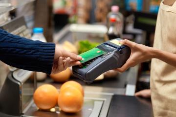 Cropped view of young adult man holding plastic credit card in hand, using terminal and paying for shopping in supermarket